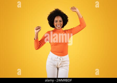 Jeune femme rayonnante aux cheveux afro lève joyeusement ses bras dans un geste de célébration Banque D'Images