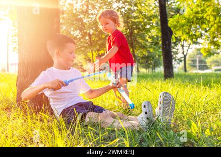 Garçon assis sous l'arbre soufflant des bulles de savon avec le tout-petit approchant Banque D'Images