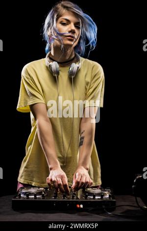Fille portant des écouteurs se concentre sur le mixage de la musique sur une table d'harmonie. DJ femme avec cheveux bleus et t-shirt jaune. Banque D'Images