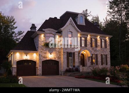 Luxueuse maison illuminée de deux étages en pierre taillée brune et bronzée avec garage pour deux voitures et allée en pierre pavée en été au crépuscule. Banque D'Images