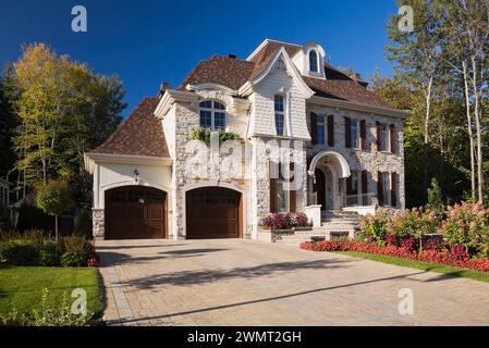 Luxueuse maison en pierre taillée brune et brune de deux étages avec garage pour deux voitures, allée en pierre pavée et cour avant paysagée en été. Banque D'Images