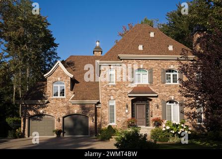 Luxueuse façade de maison de style manoir français de deux étages nuancé bronzé et brun avec garage pour deux voitures et cour avant paysagée en été. Banque D'Images