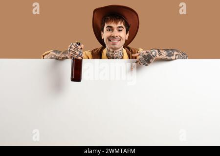 Barman masculin habillé comme un cow-boy avec une bouteille de bière pointant vers une affiche vierge sur fond beige Banque D'Images