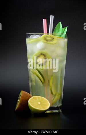 Limonade rafraîchissante avec des morceaux de fruits et de glace dans un grand verre à facettes sur fond noir, à côté d'un citron vert mûr et de kiwi. Arrangement vertical Banque D'Images