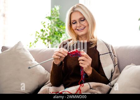 Portrait de mignonne grand-mère souriante avec des cheveux blonds portant un pull confortable lors du tricotage de chaussettes en laine faites à la main assis sur divan à l'intérieur Banque D'Images
