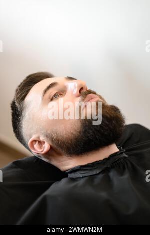 Un jeune homme avec une barbe dans une cape noire est assis sur une chaise dans un salon de coiffure. Le client attend une coupe de cheveux et le rasage de la barbe. Banque D'Images