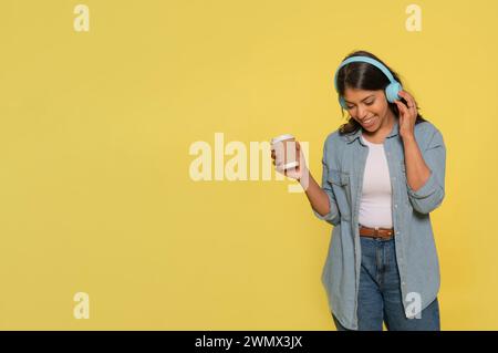 Heureuse jeune femme indienne emportée avec de la musique danse insouciante avec les bras levés porte des écouteurs stéréo sur ses oreilles habillées de jeans et un sh denim Banque D'Images