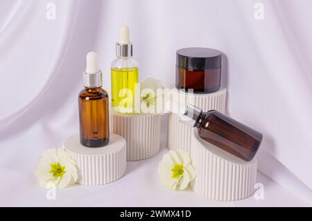 Ensemble de produits cosmétiques naturels d'auto-soin sur des podiums gaufrés blancs sur le fond de tissu de satin blanc avec des fleurs de bégonia. Vue de face Banque D'Images