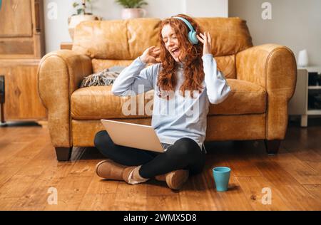 heureuse jeune dame bouclée assise sur le sol, buvant du thé, écoutant des chansons préférées sur des écouteurs, et chantant, profitant de la journée ensoleillée à la maison. Banque D'Images