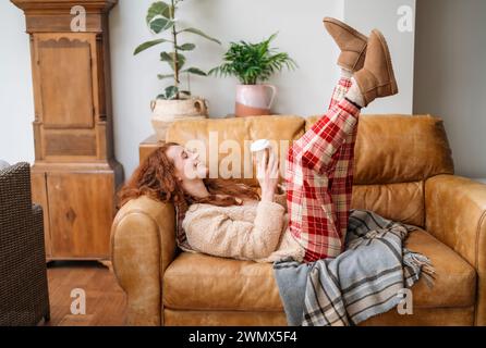 Une jeune femme en pyjama couchée sur un canapé et buvant du café dans le salon Banque D'Images