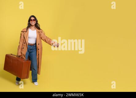 Une femme en Jean, un T-shirt blanc, et un manteau marron portant une valise et prenant un selfie par téléphone sur fond jaune. Des gens heureux qui vont sur Hol Banque D'Images