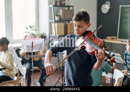 Plan moyen d'un garçon caucasien pratiquant le violon pour l'orchestre de l'école en classe, ses camarades de classe assis en arrière-plan Banque D'Images