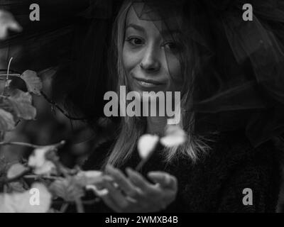 Une image horizontale calme en noir et blanc d'une jeune femme blanche tenant doucement une feuille, ornée d'un grand chapeau noir, dans une ambiance rétro de mode sombre Banque D'Images