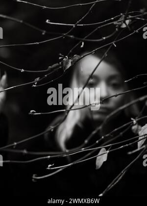 Visage floue blanc pâle d'une jeune femme partiellement obscurci par des branches dans un décor noir et blanc, mise au point sélective et effet bokeh, gothique foncé Banque D'Images