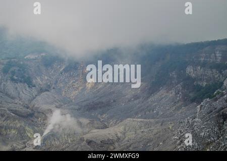 Falaises rocheuses d'un cratère volcanique brumeux avec des dépôts de cendres visibles et de la fumée montante, en Indonésie Banque D'Images