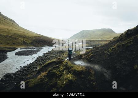 Un voyageur masculin solitaire est captivé par la sérénité d'une rivière sinueuse qui traverse les collines verdoyantes d'une vaste vallée en Islande. Banque D'Images