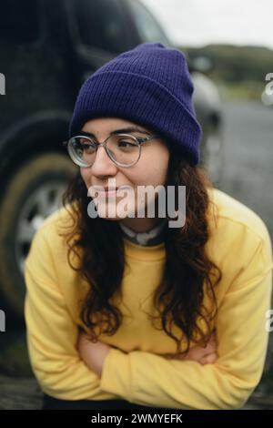 Une jeune femme réfléchie aux cheveux bouclés portant des lunettes, un pull jaune et un bonnet violet repose son menton sur son bras à l'extérieur. Banque D'Images