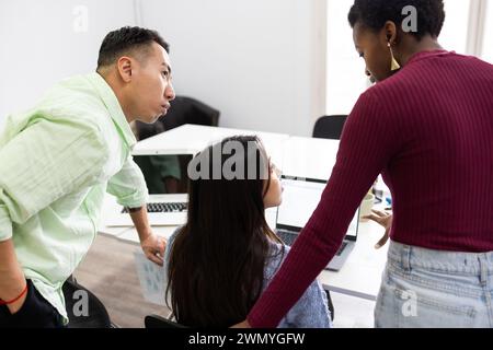 Trois professionnels divers sont engagés dans une discussion au-dessus d'un ordinateur dans un cadre de bureau lumineux, mettant en valeur le travail d'équipe et la collaboration. Banque D'Images