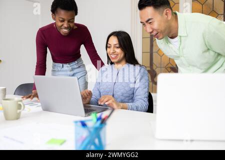 Trois collègues travaillant ensemble avec un ordinateur portable dans un environnement de bureau lumineux, partageant des idées et collaborant sur un projet. Banque D'Images
