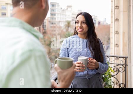 Une jeune femme sourit tout en tenant une tasse, ayant une conversation avec un partenaire sur un balcon donnant sur un paysage urbain. Banque D'Images