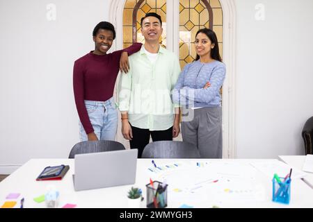 Un groupe multiethnique de trois professionnels se tient confiant dans un cadre de bureau moderne, souriant à la caméra, avec un ordinateur portable et du matériel de travail dessus Banque D'Images