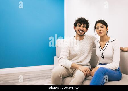 Un jeune couple s'assoit confortablement sur un canapé blanc, souriant et posant avec désinvolture contre un mur bleu dans leur maison confortable. Banque D'Images
