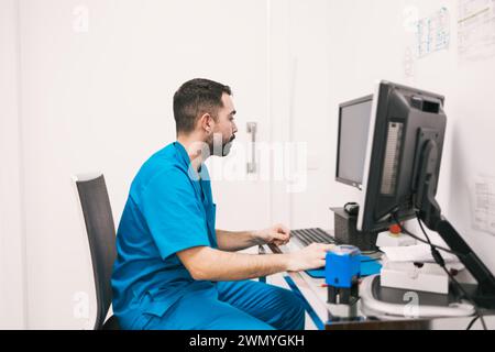 Un infirmier spécialisé dans les gommages se concentre sur la mise à jour des dossiers des patients sur un ordinateur dans une clinique médicale. Banque D'Images