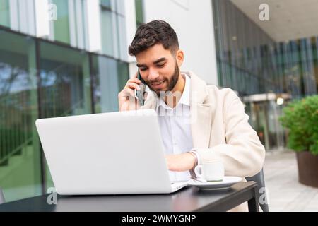 Homme d'affaires souriant s'engageant dans une conversation téléphonique tout en travaillant sur son ordinateur portable dans un coin salon extérieur d'un immeuble de bureaux moderne Banque D'Images