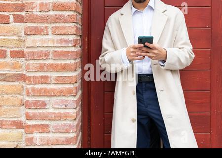 Gros plan d'un homme d'affaires anonyme dans un trench coat utilisant un smartphone contre un mur de briques texturé, incarnant le professionnalisme urbain Banque D'Images