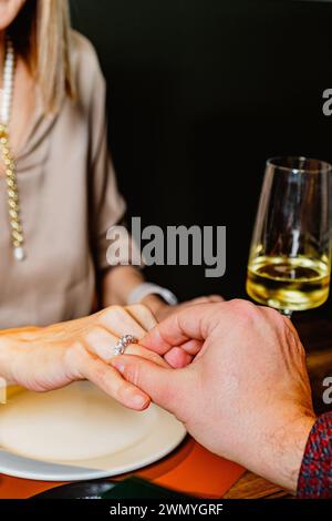 Gros plan de la main d'un homme plaçant une bague de fiançailles sur le doigt d'une femme lors d'un dîner romantique, avec un verre à vin en arrière-plan Banque D'Images