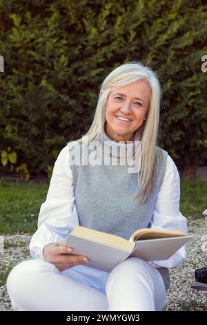Femme mature souriante assise à l'extérieur lisant un livre, avec une caméra à côté d'elle, dépeignant un style de vie actif et engagé Banque D'Images