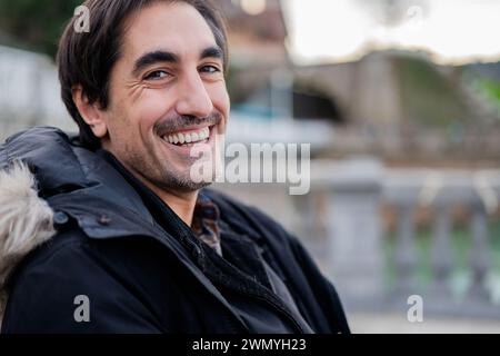 Un portrait d'un homme joyeux avec un sourire chaleureux, portant une tenue d'hiver, y compris une veste noire avec une capuche doublée de fourrure à l'extérieur Banque D'Images