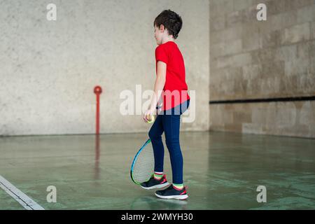 Jeune garçon dans un t-shirt rouge et un pantalon bleu tenant une raquette de tennis et une balle, prêt à jouer à l'intérieur. Banque D'Images