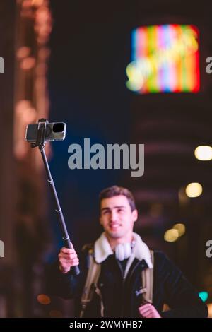 Jeune homme capturant des moments à l'aide d'un smartphone sur un bâton selfie avec des lumières colorées de la ville en arrière-plan Banque D'Images