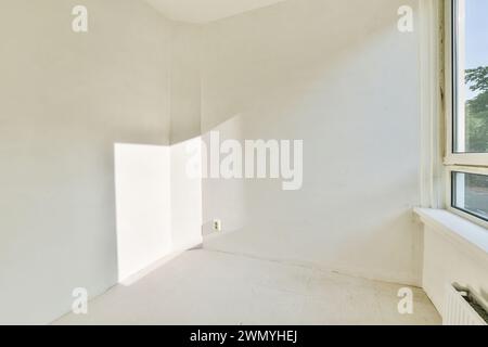 Une pièce vide avec des murs blancs, la lumière naturelle du soleil qui traverse une fenêtre et un sol clair, créant un espace minimaliste. Banque D'Images
