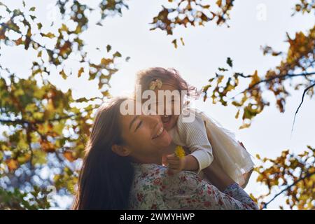 D'en bas, une mère ethnique soulève joyeusement sa jeune fille dans un parc ensoleillé, entourée de feuilles d'automne en Californie, aux États-Unis, exprimant la joie et l'amour. Banque D'Images