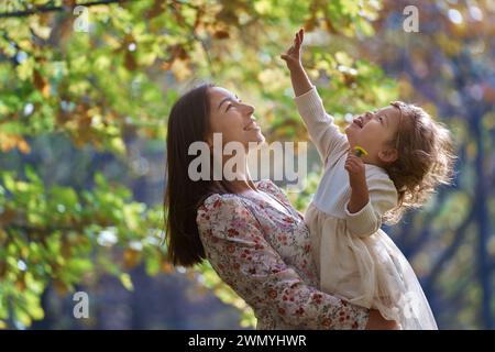 Vue de côté de la mère joyeuse soulève sa fille rigolante dans un parc ensoleillé, entouré par la nature en Californie, partageant un moment tendre ensemble. Banque D'Images