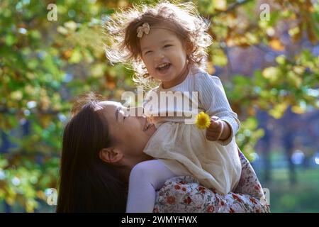 Une scène réconfortante d'une mère ethnique soulevant sa fille souriante dans un parc californien ensoleillé, entouré par la beauté de la nature. Banque D'Images