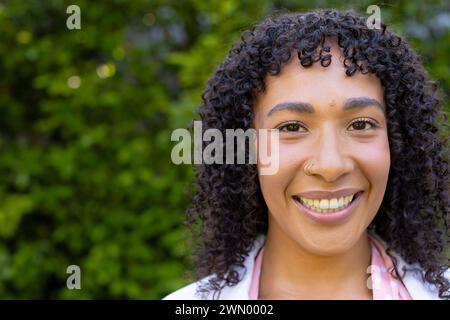 Jeune femme biraciale aux cheveux bouclés sourit brillamment à la maison, portant une écharpe rose, avec un espace de copie Banque D'Images