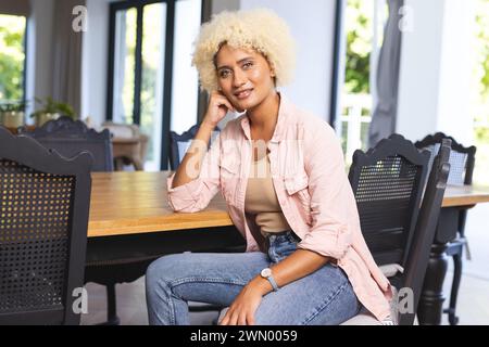 Jeune femme biraciale aux cheveux blonds bouclés est assise à une table en bois, souriant doucement Banque D'Images