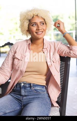 Jeune femme biraciale aux cheveux blonds bouclés s'assoit décontractée sur une chaise Banque D'Images