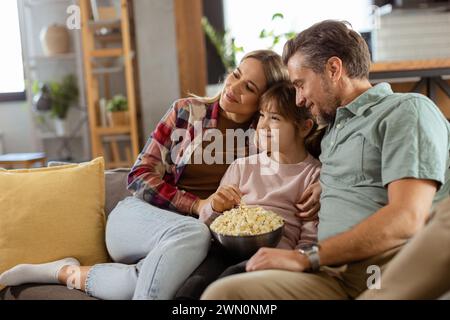 Une famille de trois est confortablement nichée sur un canapé, leurs visages reflétant l'excitation et l'attention comme ils partagent un bol de pop-corn pendant un susp Banque D'Images