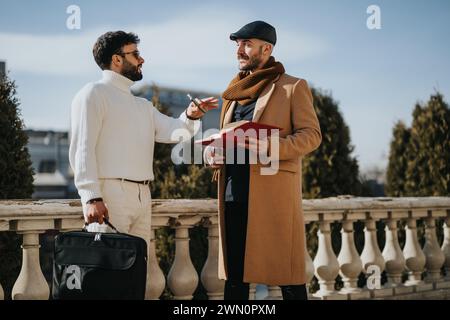 Deux hommes bien habillés s'engagent dans une conversation décontractée tenant un dossier rouge par une journée ensoleillée. Ils affichent un comportement amical dans un contexte urbain. Banque D'Images