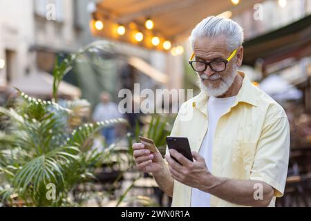 Homme aux cheveux gris senior touriste se promène dans la ville du soir, le retraité tient le téléphone et la carte de débit bancaire dans les mains, choisit des cadeaux dans la boutique en ligne, achète à distance et livre des services. Banque D'Images