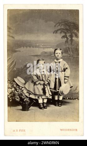 Carte de visite victorienne originale, charmante et claire (carte de visite ou CDV) de 2 jeunes enfants victoriens, garçons victoriens, frères et sœurs (pas les filles comme ont les coiffures des garçons) dans des robes assorties avec un look crinoline et des pantaloons dessous pour la tenue modérée des chapeaux de paille, détail de bouton. Le garçon plus âgé a une expression douloureuse sur son visage. Pas étonnant ! Du studio photographique d'Alfred Samuel Fisk, John's préparé Woodbridge, Suffolk, Angleterre, Royaume-Uni vers 1865., Banque D'Images