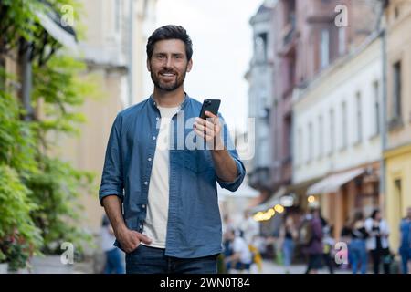 Portrait d'un jeune homme debout dans une rue de la ville, tenant une main dans sa poche et tenant un téléphone de l'autre main, souriant et regardant l'appareil photo. Banque D'Images
