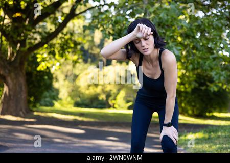 Jeune femme active en vêtements noirs de sport debout dans le parc et penchée de la fatigue après l'entraînement, la course et le marathon, essuyant la sueur avec la main du front et se reposant. Banque D'Images