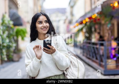 Une jeune femme indienne se promène dans les rues de la ville, voyage, utilise le téléphone avec le sourire, fouille à travers le navigateur. Banque D'Images