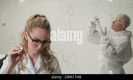 Une femme chimiste écrit une formule sur verre. Un homme caucasien âgé en costume de protection fait des tests. Banque D'Images