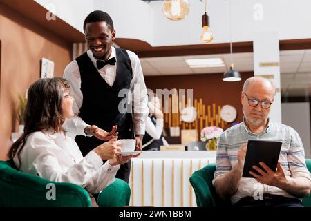 Couple de personnes âgées sur le canapé du hall de l'hôtel, en utilisant une tablette numérique et boire du café tandis que l'employé assure un séjour confortable. Les personnes âgées retraitées apprécient une tasse de thé et naviguent sur Internet dans le coin salon confortable. Banque D'Images